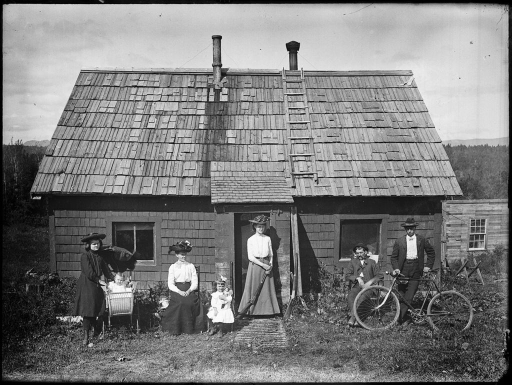 Miniature of Family posing outside their small rural home. Among them are a young man with a bicycle, a woman with a shotgun & a baby in a carriage