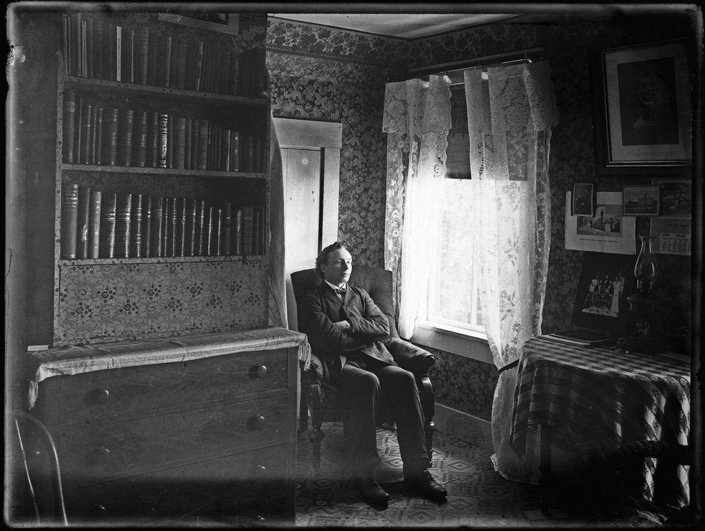 Miniature of Man seated by a window in a small room with bookcase and pictures on a wall