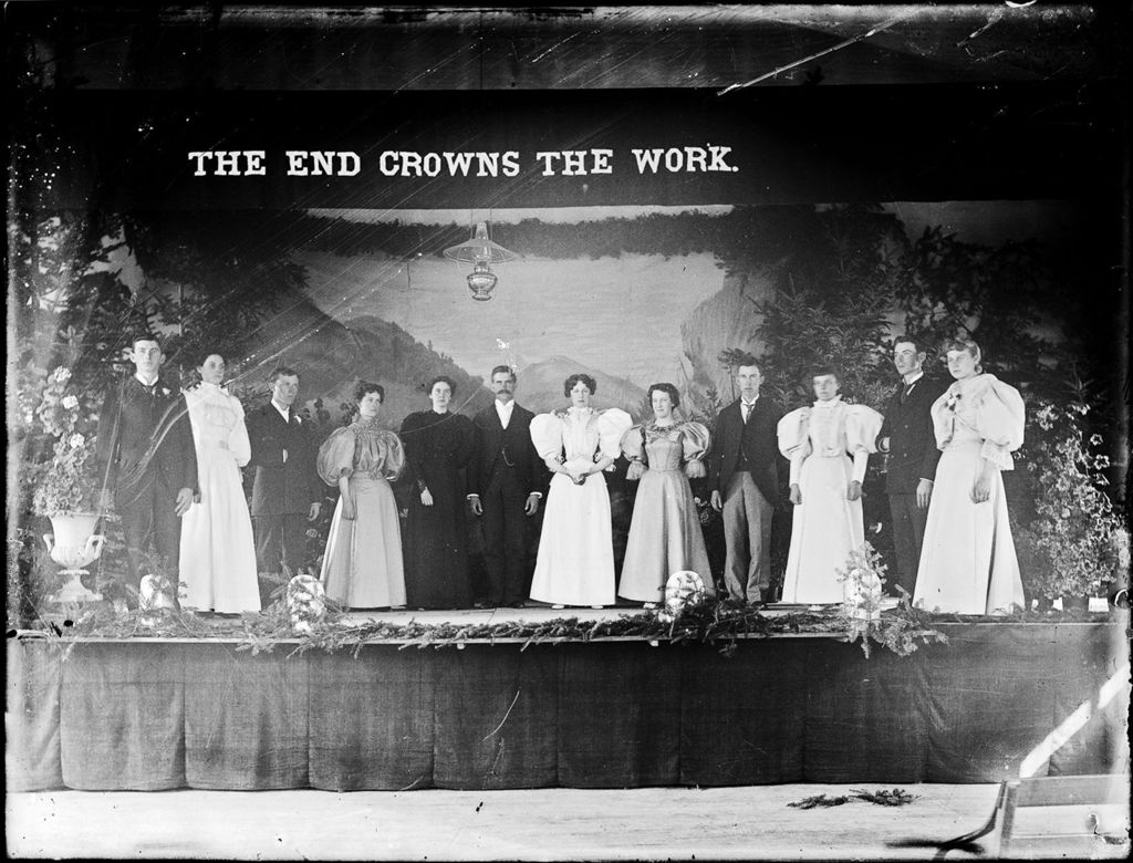 Miniature of Group of well dressed men and women standing on a stage under a banner that reads "The End Crowns the Work"